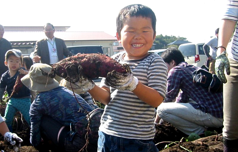 イモ畑で、収穫したサツマイモを持って笑う子どもの写真
